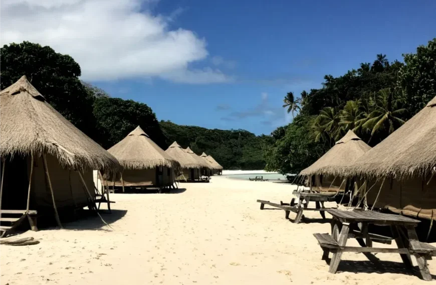 Strohgedeckte Hütten säumen einen Sandstrand, der unter einem klaren blauen Himmel zu einem tropischen Wald im Südsee Camp führt.