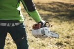 Eine Person in einer grünen Jacke, die im Freien eine Stihl-Kettensäge in der Hand hält, im Hintergrund sind Gras und Sonnenlicht zu sehen.